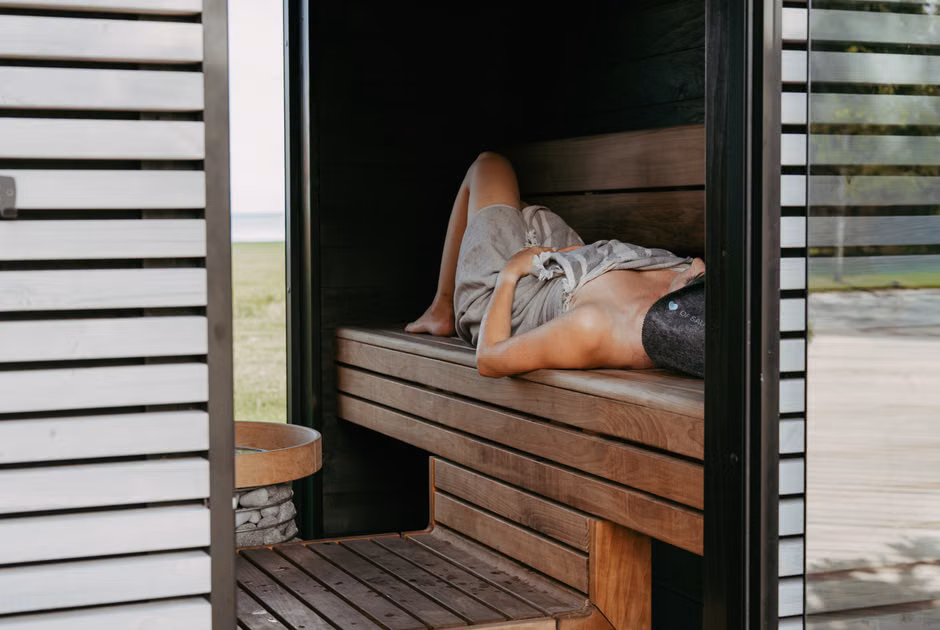 Wellness Facility Standards: International Quality Benchmarks for Spas Person relaxing inside a modern sauna with Nordic design, lying with a towel and sauna hat for wellness.